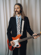 Tropicana Tie in white silk with brown and blue paisley, worn with a black jacket and light blue shirt, three-quarter view holding a red electric guitar