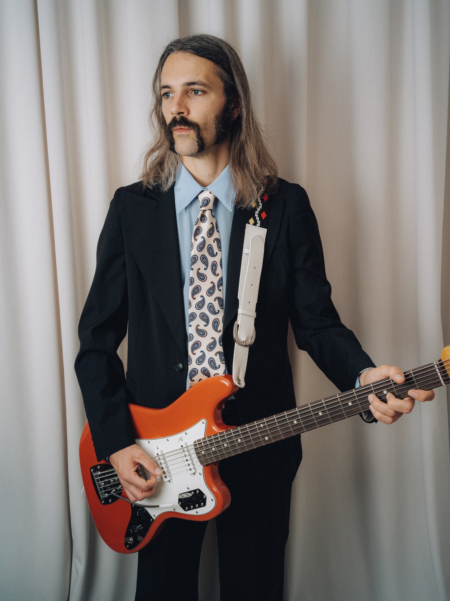 Tropicana Tie in white silk with brown and blue paisley, worn with a black jacket and light blue shirt, three-quarter view holding a red electric guitar