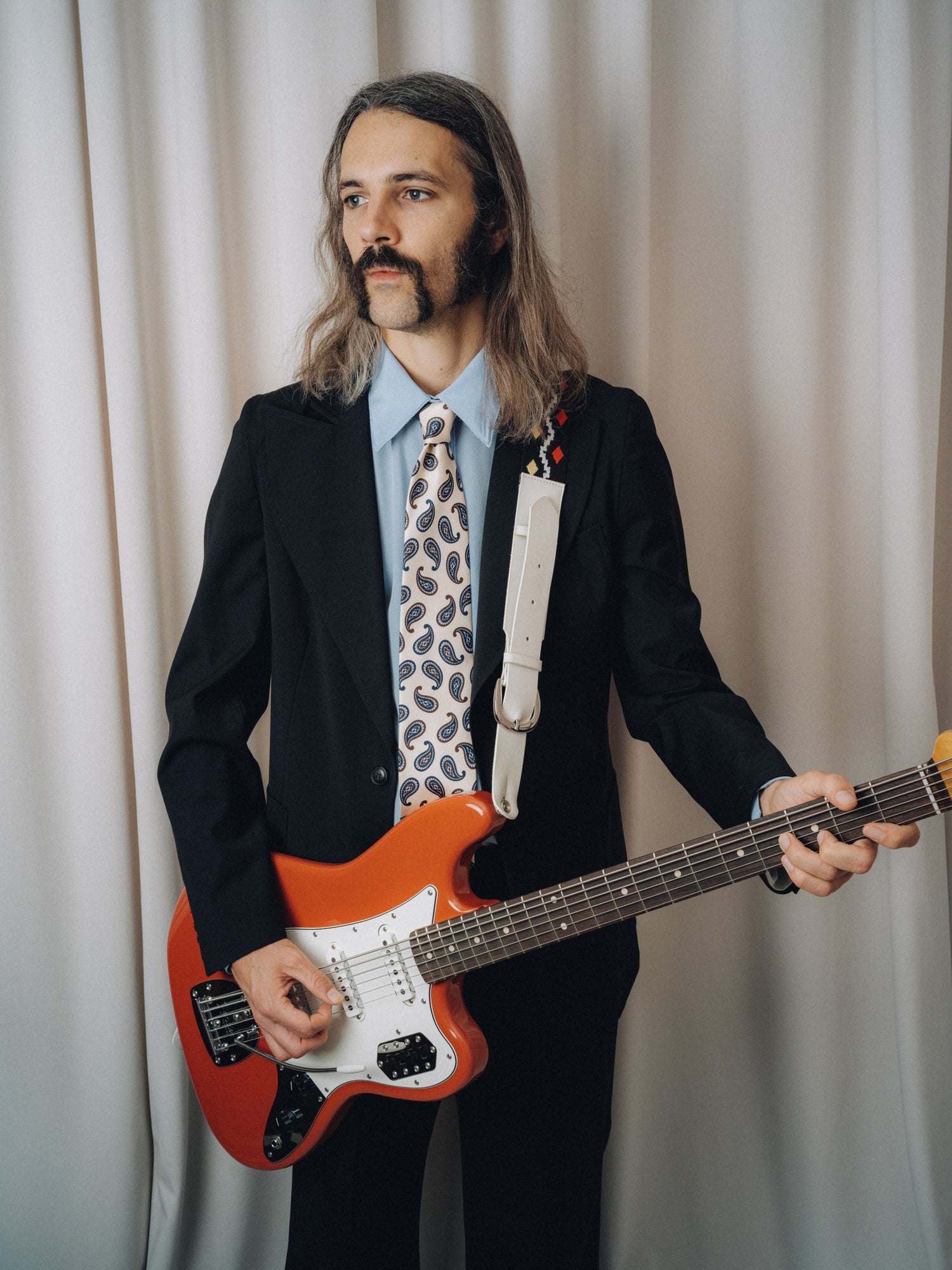 Tropicana Tie in white silk with brown and blue paisley, worn with a black jacket and light blue shirt, three-quarter view holding a red electric guitar