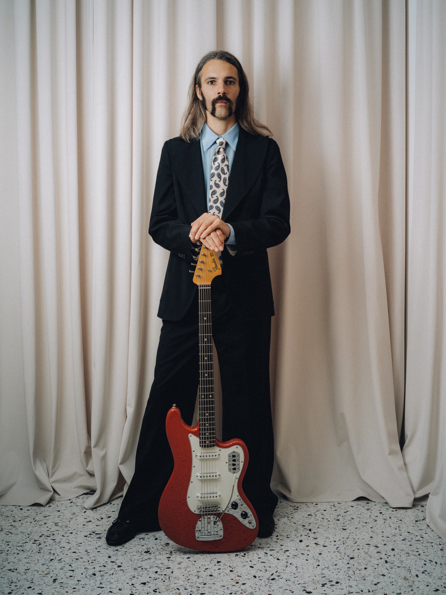 Tropicana Tie in white silk with brown and blue paisley, full figure with the tie centred, holding a red electric guitar