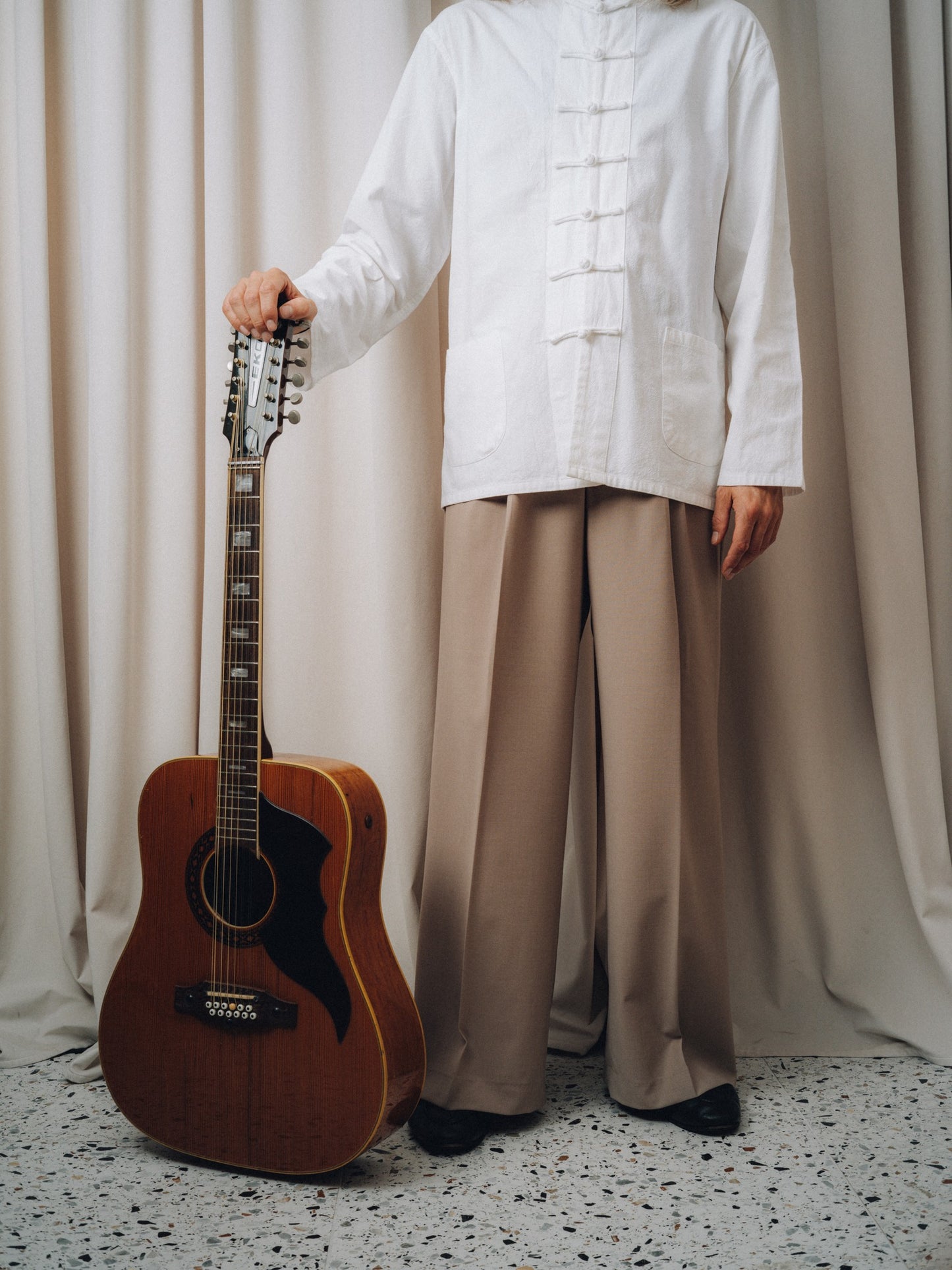 Progressiva Shirt in white with knot buttons, worn with taupe trousers and a twelve-string acoustic guitar